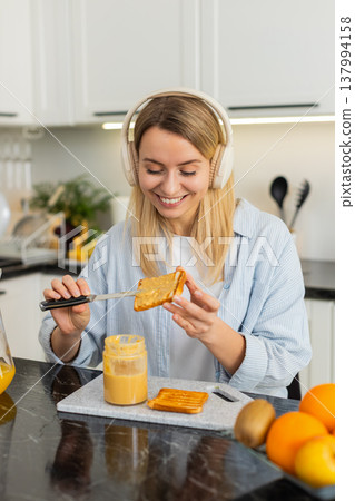 Young woman spreading peanut butter on toast while listening to music with headphones in own kitchen Young woman spreading peanut butter on toast while listening to music with headphones in own kitchen 137994158