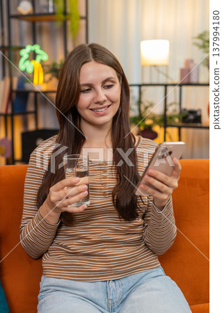 Young woman drinks water from glass plans tasks on smartphone focused calm and replies to messages 137994180