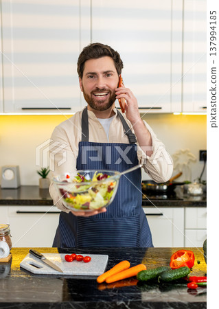 Middle-aged man tasting fresh vegetable salad while talking on smartphone in home kitchen for diet Middle-aged man tasting fresh vegetable salad while talking on smartphone in home kitchen for diet 137994185
