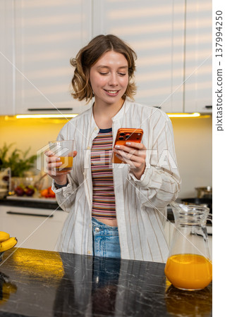Young woman using smartphone while drinking orange juice in kitchen during healthy breakfast break Young woman using smartphone while drinking orange juice in kitchen during healthy breakfast break 137994250