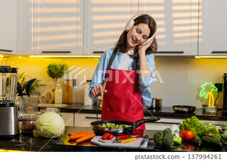 Young woman dancing to music while frying vegetables in kitchen for healthy diet cooking meal prep Young woman dancing to music while frying vegetables in kitchen for healthy diet cooking meal prep 137994261
