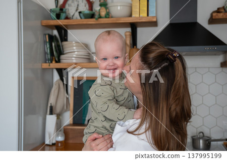Mother holds smiling baby in kitchen interior with natural light and home decor, close interaction and eye contact in everyday domestic setting. Family bonding, motherhood, parenting, lifestyle 137995199