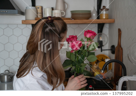 Woman holds bouquet of pink roses in kitchen interior with natural light and home decor, everyday moment of care, attention in domestic setting. Family care, lifestyle content, home routine, wellbeing 137995202