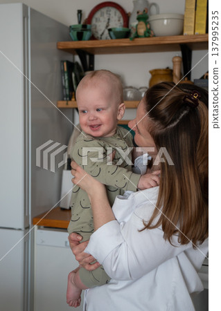 Mother holds smiling baby in kitchen interior with natural light and shelves with home items, warm interaction and everyday domestic moment. Family bonding, motherhood, parenting content 137995235