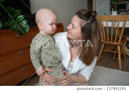 Mother holds baby at home interior with warm light and wooden furniture, close interaction and eye contact between parent and child in everyday setting. Family bonding, motherhood, parenting 137995295
