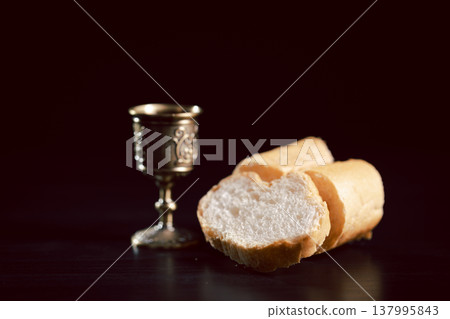 Communion still life with bread on a wooden plate, red wine in a small cup and ornate chalice beside a Holy Bible on dark table, symbolizing Eucharist, worship, faith and remembrance with copy space. 137995843