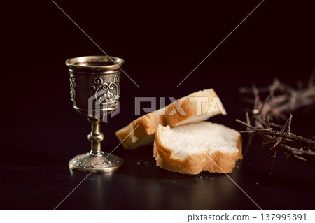 Moody still life of ornate chalice with red wine and sliced bread beside a crown of thorns on dark background, symbolizing Christian communion, sacrifice, worship and faith with copy space. Moody still life of ornate chalice with red wine and sliced bread beside a crown of thorns on dark background, symbolizing Christian communion, sacrifice, worship and faith with copy space. 137995891