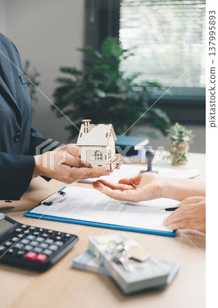 Real estate agent handing wooden house model to client over property contract at office desk, concept of home purchase, mortgage approval, ownership transfer, key handover and investment. 137995893