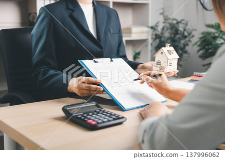 Real estate agent presenting property contract and wooden house model to client at office desk, concept of home purchase, mortgage planning, ownership transfer, consultation and closing deal. Real estate agent presenting property contract and wooden house model to client at office desk, concept of home purchase, mortgage planning, ownership transfer, consultation and closing deal. 137996062
