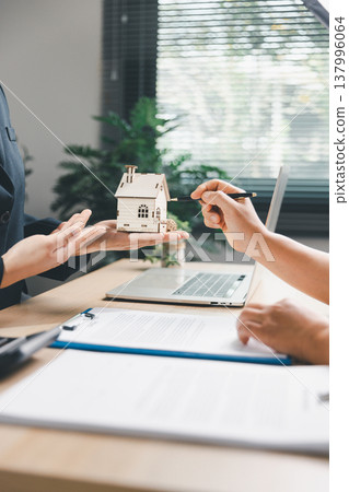 Real estate agent presenting wooden house model to client pointing during property consultation at office desk with laptop and contract, concept of home purchase, mortgage planning and ownership. Real estate agent presenting wooden house model to client pointing during property consultation at office desk with laptop and contract, concept of home purchase, mortgage planning and ownership. 137996064