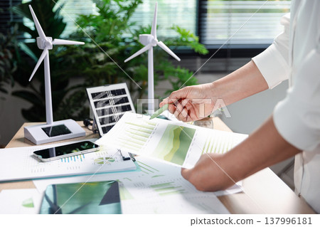 Sustainability analyst points to green ESG charts on paper report beside wind turbines and solar panel model, reviewing renewable energy metrics and carbon reduction plan in bright office, copy space. Sustainability analyst points to green ESG charts on paper report beside wind turbines and solar panel model, reviewing renewable energy metrics and carbon reduction plan in bright office, copy space. 137996181