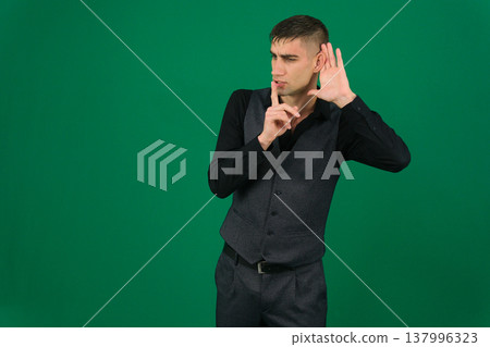 Asian man in denim jacket making silence gesture with finger on lips and hand cupped to ear, listening carefully, serious expression, concept of secret, hush, or confidential talk Asian man in denim jacket making silence gesture with finger on lips and hand cupped to ear, listening carefully, serious expression, concept of secret, hush, or confidential talk 137996323