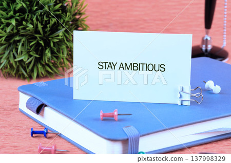White card shows STAY AMBITIOUS resting on a serene blue journal. Bright pins and a verdant plant adorn the warm wooden desk, encouraging aspirations White card shows STAY AMBITIOUS resting on a serene blue journal. Bright pins and a verdant plant adorn the warm wooden desk, encouraging aspirations 137998329
