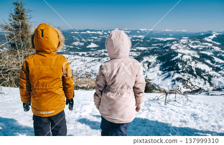 Kids standing on top of cliff in winter mountains at sunset and enjoying view of nature Kids standing on top of cliff in winter mountains at sunset and enjoying view of nature 137999310