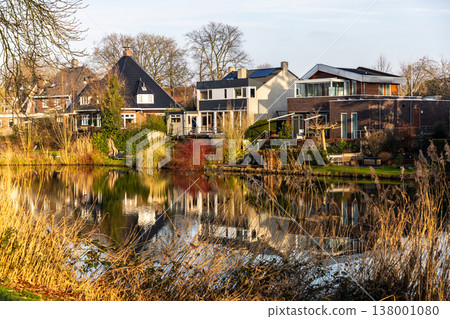 Modern and traditional suburban houses reflected in a calm pond at golden hour. Serene lakeside neighborhood with autumn reeds, sunlit architecture, and peaceful atmosphere in Europe. 138001080