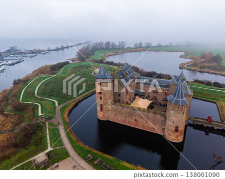 High aerial view of the historic Muiderslot Castle, a perfectly preserved medieval brick fortress near Amsterdam, Netherlands, surrounded by a dark moat and a network of water channels on a misty day. High aerial view of the historic Muiderslot Castle, a perfectly preserved medieval brick fortress near Amsterdam, Netherlands, surrounded by a dark moat and a network of water channels on a misty day. 138001090