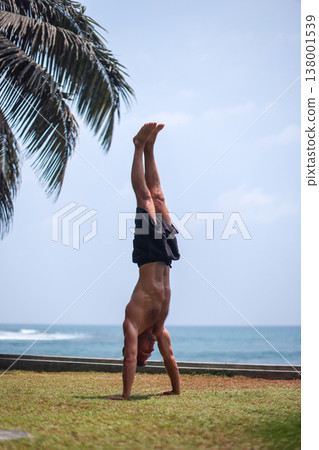 Man holding handstand on grass near ocean in tropical setting Man holding handstand on grass near ocean in tropical setting 138001539
