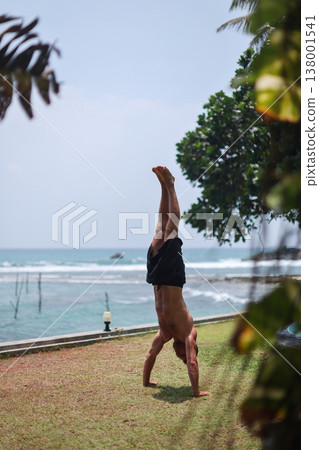 Man holding handstand on grass near ocean in tropical setting Man holding handstand on grass near ocean in tropical setting 138001541