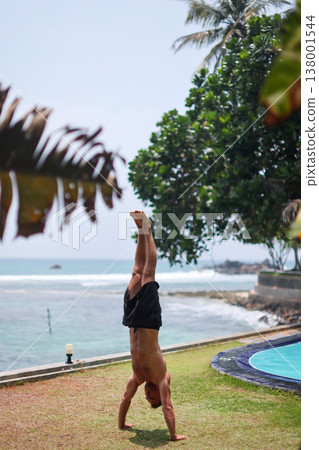 Man holding handstand on grass near ocean in tropical setting Man holding handstand on grass near ocean in tropical setting 138001544