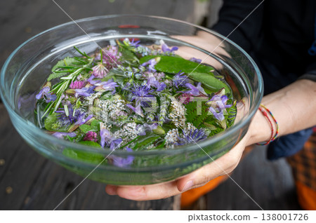 Hands hold a clear glass bowl filled with water, fresh purple wildflowers, green herbs and foliage, creating a natural flower bath for wellness, relaxation, and herbal medicine Hands hold a clear glass bowl filled with water, fresh purple wildflowers, green herbs and foliage, creating a natural flower bath for wellness, relaxation, and herbal medicine 138001726