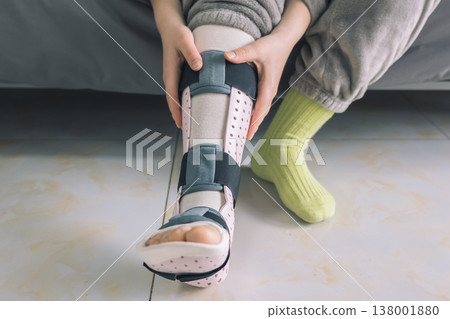 A person's hands are carefully adjusting the black straps of a pink perforated medical leg brace on their lower left leg. 138001880