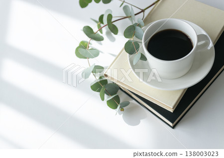 一杯盛在白色杯子裡的熱咖啡，一本書，還有幾朵粉紅色的花。陽光灑進房間，享受愜意的清晨時光。留有文字空間。 138003023