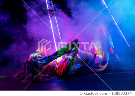 Woman in glowing UV body paint lying in smoky, futuristic neon light. Woman in glowing UV body paint lying in smoky, futuristic neon light. 138003374