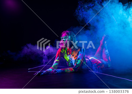 Woman covered in neon UV body paint with glowing glasses lies in colorful smoke. Woman covered in neon UV body paint with glowing glasses lies in colorful smoke. 138003376