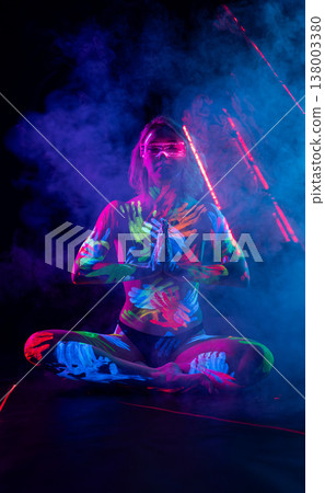 Woman in UV body paint and cyber glasses sits in a meditative pose amidst purple smoke and laser beams. Woman in UV body paint and cyber glasses sits in a meditative pose amidst purple smoke and laser beams. 138003380