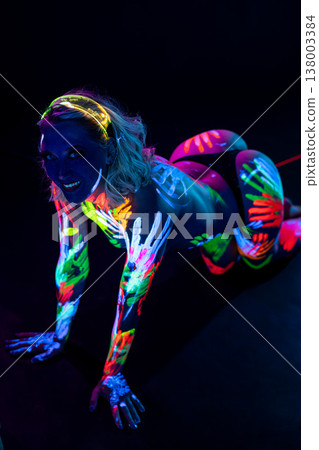 Woman with vibrant neon body paint handprints glowing under blacklight on dark background. 138003384