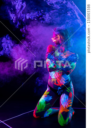 Woman with glowing neon handprints and futuristic glasses kneeling in blacklight on a dark background. 138003386