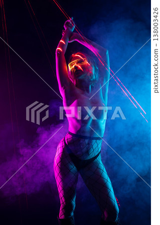 Sensual cyberpunk woman in glowing glasses posing with neon pink and blue laser beams in a dark, smoky studio. Sensual cyberpunk woman in glowing glasses posing with neon pink and blue laser beams in a dark, smoky studio. 138003426