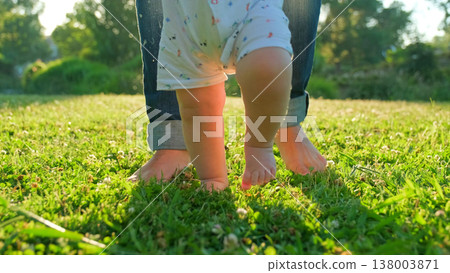 Child learns to take the first steps on the grass. Baby learns to walk with the help of his mother on a green grass in the park. Walking children's bare feet on a green lawn close-up. Child learns to take the first steps on the grass. Baby learns to walk with the help of his mother on a green grass in the park. Walking children's bare feet on a green lawn close-up. 138003871