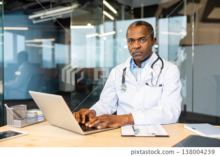 Male doctor wearing a white lab coat and stethoscope, looking directly at the camera while typing on a laptop, providing healthcare services in a professional office setting 138004239