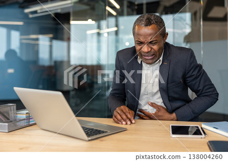 Mature businessman seated at his office desk holding his abdomen and grimacing in pain, appearing unwell and stressed while working on a laptop in a corporate workplace 138004260