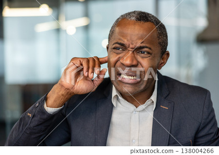 Mature man grimacing, inserting finger into his ear, indicating an irritating noise, a sudden loud sound, or experiencing an earache, tinnitus, or hearing issues at work 138004266
