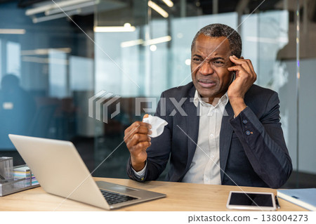Mature professional man at his office desk rubbing his temple and holding a crumpled tissue, looking unwell and fatigued from headache, stress or flu while working on a laptop Mature professional man at his office desk rubbing his temple and holding a crumpled tissue, looking unwell and fatigued from headache, stress or flu while working on a laptop 138004273