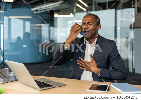 Mature businessman experiencing a respiratory attack, using an inhaler to alleviate breathing difficulties, placing a hand on his chest in an office setting 138004275