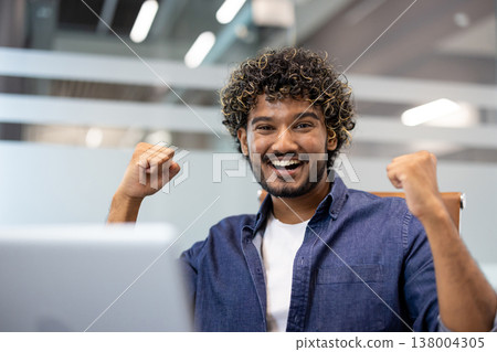 Close-up portrait of a young Indian man sitting in an office with a laptop and looking at the camera. He raised his hands up, showing a sign of strength. Close-up portrait of a young Indian man sitting in an office with a laptop and looking at the camera. He raised his hands up, showing a sign of strength. 138004305