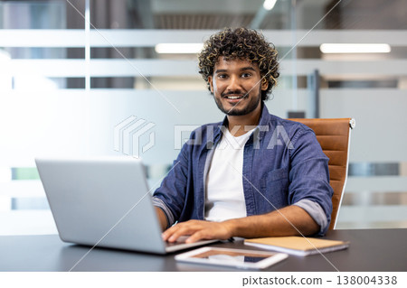 Portrait of a young Indian man sitting at a desk in the office, working on a laptop and smiling, looking at the camera. 138004338
