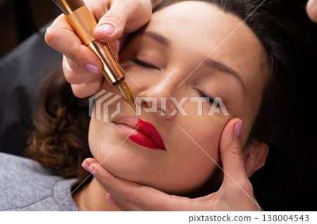 Young woman making lips tatouage in a dressing makeup room. Lips tatouage process .Woman making lips blushing. Make-up artist in beauty studio doing makeup for beautiful girl. Making mua. 138004543
