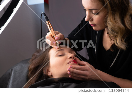 Young woman making tatouage for eyebrows in a dressing makeup room. Eyebrows make up process. Make-up artist in beauty studio doing makeup for beautiful girl. Making mua. 138004544