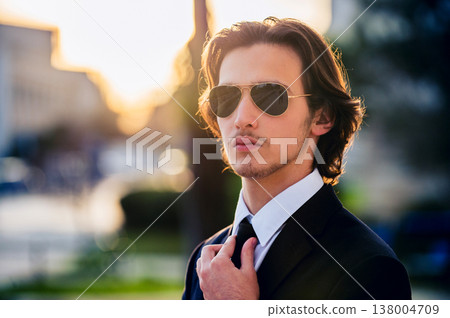 Closeup portrait of a male fashion model with sunglasses in a stylish blue suit with tie. Handsome young man model in elegant business suit poses with sunglasses with bright sunlight on background. Closeup portrait of a male fashion model with sunglasses in a stylish blue suit with tie. Handsome young man model in elegant business suit poses with sunglasses with bright sunlight on background. 138004709