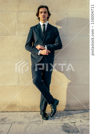 Handsome young man model in a blue elegant business suit poses against a light stone wall outdoors in summer. Male Fashion model poses in front of the camera in a stylish blue suit and white shirt. Handsome young man model in a blue elegant business suit poses against a light stone wall outdoors in summer. Male Fashion model poses in front of the camera in a stylish blue suit and white shirt. 138004711