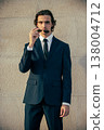 Handsome young man model in a blue elegant business suit poses with sunglasses against a light stone wall outdoors in summer. Male Fashion model poses in a stylish blue suit and white shirt. 138004712