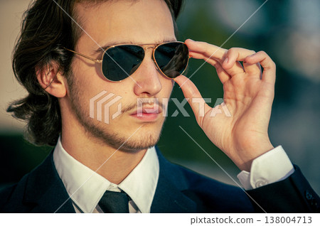 Closeup portrait of a male fashion model with sunglasses in a stylish blue suit with tie. Handsome young man model in elegant business suit poses with sunglasses. Closeup portrait of a male fashion model with sunglasses in a stylish blue suit with tie. Handsome young man model in elegant business suit poses with sunglasses. 138004713