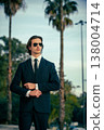 Stylish portrait of  a male fashion model with sunglasses in a elegance blue suit with tie. Handsome young man model in modern business suit poses with sunglasses posing in the street. 138004714