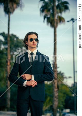 Stylish portrait of a male fashion model with sunglasses in a elegance blue suit with tie. Handsome young man model in modern business suit poses with sunglasses posing in the street. Stylish portrait of a male fashion model with sunglasses in a elegance blue suit with tie. Handsome young man model in modern business suit poses with sunglasses posing in the street. 138004714