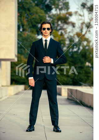 Stylish portrait of  a male fashion model with sunglasses in a elegance blue suit with tie. Handsome young man model in modern business suit poses with sunglasses posing in the street. 138004715