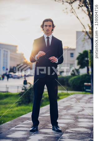 Portrait of a male fashion model in a stylish blue suit with tie posing in the city. Handsome young man model in elegant business suit poses outdoors with bright sunlight on background. Portrait of a male fashion model in a stylish blue suit with tie posing in the city. Handsome young man model in elegant business suit poses outdoors with bright sunlight on background. 138004716